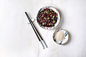 Korean-style vegan beetroot salad. Marinated grated beetroot with sesame seeds and coriander leaves in a ceramic bowl. Next to it, bowl with sesame seeds and chopsticks on a white marble base. View from above