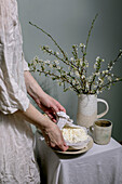 Frau in weißer Leinenkleidung serviert frischen hausgemachten Hüttenkäse, der in ein Tuch eingewickelt ist. Keramikkrug mit blühenden weißen Frühlingszweigen und Teetasse auf weißem Tischtuch. Rustikal-elegante Umgebung.