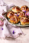 Traditional hot cross buns on a ceramic plate decorated with edible flowers. Homemade Easter buns on a light-coloured background with linen napkin and purple petals. Close-up