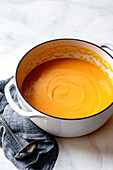 A creamy carrot soup is presented in a white pot, accompanied by a silver ladle and a dark grey cloth napkin.