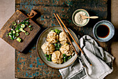 Homemade Asian steamed dumplings in a grey plate, topped with sesame seeds and green onions. Soy sauce, chopsticks, grey napkin nearby on a rustic wooden board, beige textured background.