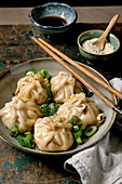 Homemade Asian steamed dumplings in a grey plate, topped with sesame seeds and green onions. Soy sauce, chopsticks, grey napkin nearby on a rustic wooden board, beige textured background.