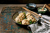 Homemade Asian steamed dumplings in a grey plate, topped with sesame seeds and green onions. Soy sauce, chopsticks, grey napkin nearby on a rustic wooden board, beige textured background.