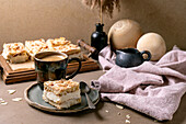 Homemade Norwegian cake Verdens Beste Kake with pudding, meringue and almonds, cut into squares, served on a wooden tray. Beige-coloured table with coffee cup, spoon, milk jug, dry reed in a bottle, beige-coloured wall.