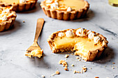 A bitten lemon tart with cream and crumble lies on a marble plate with a fork next to it.