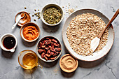 A selection of ingredients for a muesli recipe, including rolled oats, pecans, pumpkin puree, maple syrup, pumpkin seeds and spices, arranged on a marble surface.