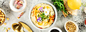 Eine schön gestaltete Schale mit cremigem Hummus, garniert mit Olivenöl, schwarzem Sesam, Pinienkernen, Mikrogrün, Paprika und essbaren Blüten, serviert mit Fladenbrot und frischen Zutaten. Perfekt als Bannerbild für lebensmittelbezogene Inhalte.