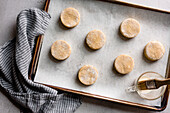 Top view of a baking tray with unbaked round biscuits on parchment paper, next to it a brush and a cup with liquid for glazing.