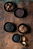 Set of five black spices: black fermented garlic, black Himalayan salt, black mustard seeds, black cardamom and fermented campota pepper in black ceramic bowls. Dark wooden background. Close-up