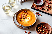 A bowl of creamy carrot soup, garnished with sliced peppers and crispy snacks, accompanied by two glasses of sparkling drink.