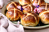 Traditional hot cross buns on a ceramic plate decorated with edible flowers. Homemade Easter buns on a light-coloured background with linen napkin and purple petals. Close-up