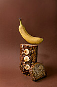 Modern still life with homemade banana bread. Loaf with walnuts, drizzled caramel, topped with fresh banana on brown background. Floating banana on top. Concept for bakery-restaurant advertising.