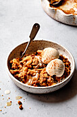 A bowl of apple crumble and two scoops of ice cream, garnished with cinnamon, on a lightly textured surface.