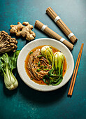 Bowl of soba noodles in a spicy broth with baby bok choy, surrounded by fresh mushrooms and ginger