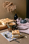 Homemade Norwegian cake Verdens Beste Kake with pudding, meringue and almonds, cut into squares, served on a wooden tray. Beige-coloured table with coffee cup, spoon, milk jug, dry reed in a bottle, beige-coloured wall.