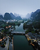 Aerial view of serene karst mountains and tranquil river with a picturesque bridge and cityscape at dusk, Guilin, China.