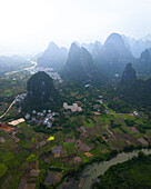 Luftaufnahme von üppig grünen Karstbergen und malerischen Feldern mit einem Fluss, der sich durch ein ländliches Dorf schlängelt, Guilin, China