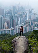 Luftaufnahme einer Frau, die auf einem Felsen steht und die atemberaubende Skyline der modernen Wolkenkratzer betrachtet, Eastern, Hongkong