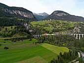 Aerial view of majestic mountains and a serene valley with a picturesque bridge amidst lush greenery, Alvaneu Bad, Switzerland.