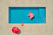 Luftaufnahme eines jungen Mädchens mit Sonnenhut am Swimmingpool mit rotem Sonnenschirm und aufblasbarem Flamingo, Sumartin, Insel Brac, Kroatien.