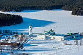 Aerial view of a serene and picturesque frozen lake surrounded by a snowy forest and a historic monastery, Leningrad Oblast, Russia.