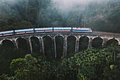 Aerial view of blue train going through famous Nine Arch railway bridge standing in lush green jungle landscape near Ella, Sri Lanka.