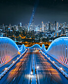 Aerial view of two people standing on a modern bridge with Dubai skyline in background at night, Dubai, United Arab Emirates-.