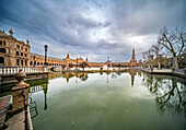 Besucher bewundern die atemberaubende Architektur der Plaza de España in Sevilla, die sich im ruhigen Wasser unter einem bewölkten Himmel spiegelt.