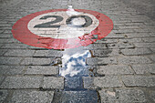 A speed limit sign marked as 20 is painted on the cobblestone pavement amidst reflections of clouds, capturing urban details.