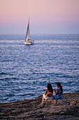 Menschen genießen die Aussicht auf den Sonnenuntergang am Pendma Chiatt (Pietra Piatta) mit einer Segelyacht im Hintergrund, Polignano a Mare, Apulien, Italien