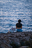 Junge Frau hört Musik und genießt den Blick auf den Sonnenuntergang von Pendma Chiatt (Pietra Piatta), Blick von hinten, Polignano a Mare, Apulien, Italien