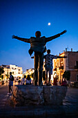 Silhouette for a man posing for a photo emulating the Statue of Domenico Modugno in Polignano a Mare at night with the moon in the background, Puglia, Italy
