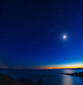 The waxing 3.5-day-old crescent Moon, with Mars and Jupiter, all amid the stars of winter shine in the darkening twilight over the Bay of Fundy at Cape Enrage near Alma, New Brunswick. This night the Moon was close to the Pleiades star cluster which appears just below the Moon. The Moon had occulted the Pleiades a few hours earlier in the evening sky over Europe. The sky is dark enought to be star-filled but retains the deep blue of twilight and the horizon colours of sunset, in this late blue-hour scene.