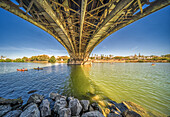 The view under Triana Bridge showcases its metal structure with the Guadalquivir River below and kayakers on the water.