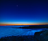 The waxing 3.5-day-old crescent Moon shines in the evening twilight at sunset over the Bay of Fundy at Cape Enrage near Alma, New Brunswick. This night the Moon was close to the Pleiades star cluster which appears just below the Moon. The Moon had occulted the Pleiades a few hours earlier in the evening sky over Europe. The stars are just beginning to appear in this blue-hour image.