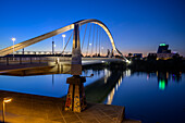 Barqueta Bridge stands gracefully against the evening sky in Seville, reflecting softly in the water below as lights begin to glow.