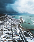 Aerial view of a snowy coastal town nestled between dark, stormy skies and the turquoise sea, Izumozaki, Niigata, Japan.