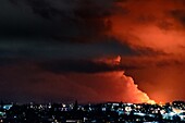 View of fiery orange clouds engulfing the night sky above a town with glowing lights, creating a dramatic contrast between darkness and radiant illumination, Grindavik, Grindavíkurbær, Iceland.