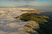 Luftaufnahme der üppig grünen Berggipfel, die ein weißes Wolkenmeer durchstoßen, mit der fernen Silhouette des Teide, La Gomera, Kanarische Inseln, Spanien.