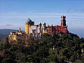 Sintra, Portugal - 11 June 2024: Aerial view of Pena Palace atop a verdant hill, its vibrant yellow, red, and grey walls contrasting with the deep green forest below.