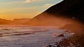 Aerial view of sunset over rugged coastline with waves and mountains, Scarborough, Western Cape, South Africa.