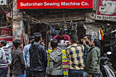 New Delhi, India - 24 June 2025: View of locals gathered around a street vendor's stall under the vibrant red sign of 'Sudarshan Sewing Machine Co.', a scene of bustling urban life.