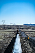 View of a silvery pipeline stretching across the arid landscape, paralleling a road under a clear blue sky, Nesjavellir, Grímsnes- og Grafningshreppur, Iceland.