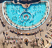 Aerial view of the vibrant Szechenyi thermal bath complex, where turquoise waters meet sun-soaked relaxation areas, creating a lively contrast of cool blues and warm earth tones, Budapest, Hungary.