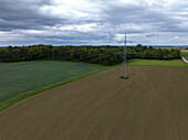 Aerial view of a stark electricity pylon standing amidst the contrasting textures of freshly tilled fields and verdant forest under a brooding sky, Southwest Germany, Baden-Württemberg, Germany.