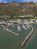 Luftaufnahme des Hafens, eingebettet zwischen dem türkisfarbenen Meer und schroffen Bergen, ein Kontrast zwischen der rauen Schönheit der Natur und den von Menschenhand geschaffenen Strukturen, Gordans Bay, Kapstadt, Westkap, Südafrika.