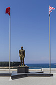 View of a bronze statue stands proudly between the flags of Turkey and Northern Cyprus against the backdrop of the azure sea, Girne, Northern Cyprus.