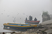 New Delhi, India - 24 June 2025: View of boats resting on the shore, as figures gather amidst the ethereal mist, birds soaring above the water, creating a tranquil yet captivating scene.