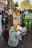 Jaipur, Indien - 24. Juni 2025: Blick auf eine Frau in einem gelben Schal, die in der Nähe eines Baumes steht, während ein Kind mit einem anderen Kind am Straßenrand Essen teilt.