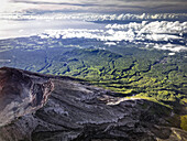 Luftaufnahme eines zerklüfteten Vulkangipfels, der in eine üppige, grüne Landschaft abfällt und unter einem wolkenverhangenen Himmel auf den weiten Ozean trifft, Vulkan Agung, Karangasem, Indonesien.